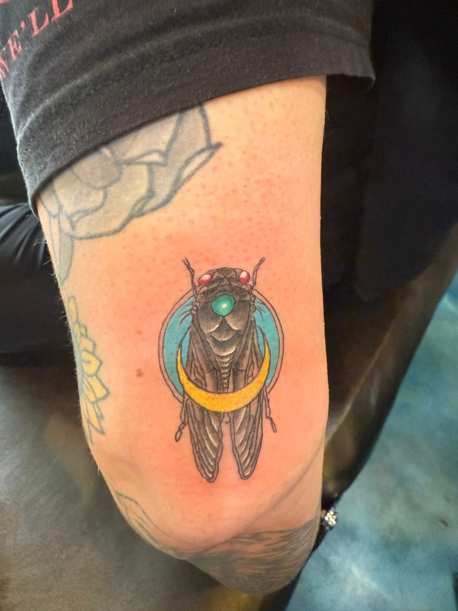 A tattoo on an arm featuring a detailed cicada with red eyes and a turquoise circle, accented by a gold crescent moon.