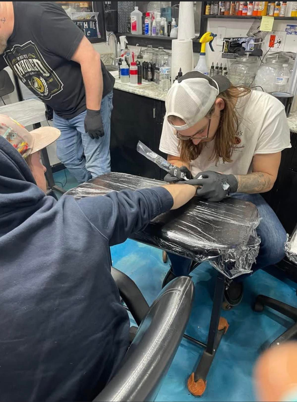 A tattoo artist in a cap and gloves works on a person's forearm in a professional tattoo studio.