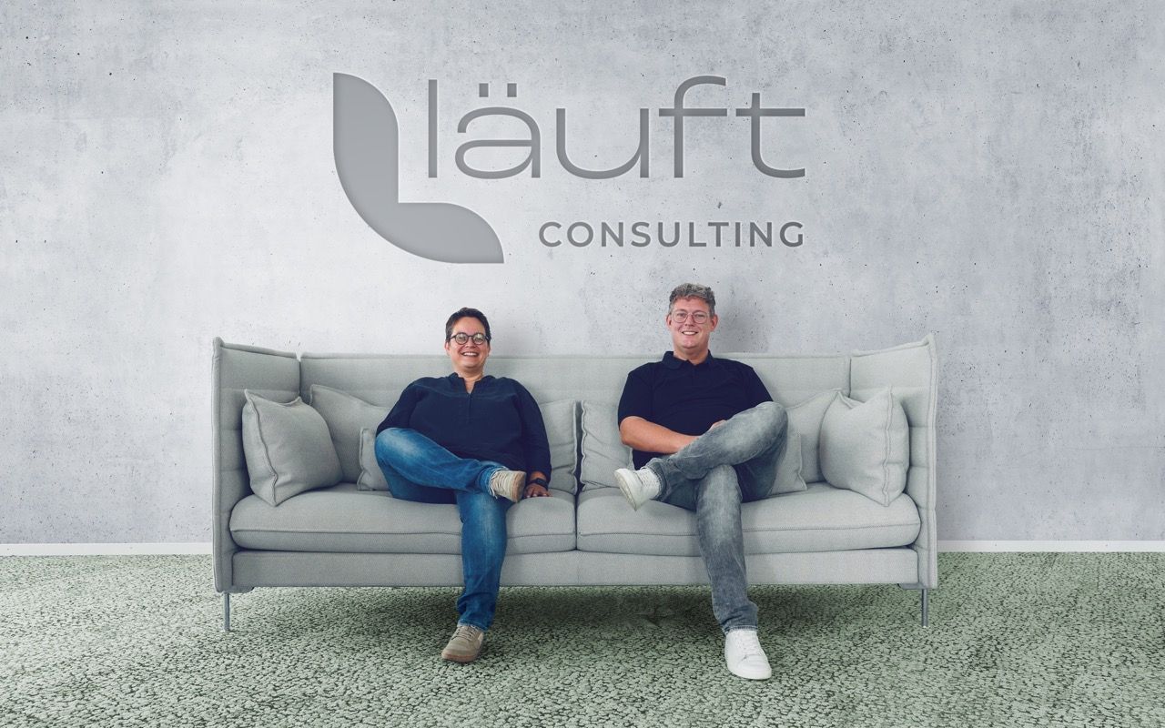 Elke und Jens mit übergeschlagenen Beinen lächelnd auf einer Vitra Couch vor einer Betonwand mit dem Logo von läuft Consulting