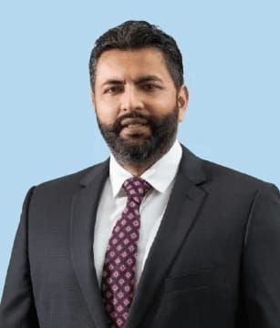 Man in suit with a beard, looking towards the camera, against a light blue background.
