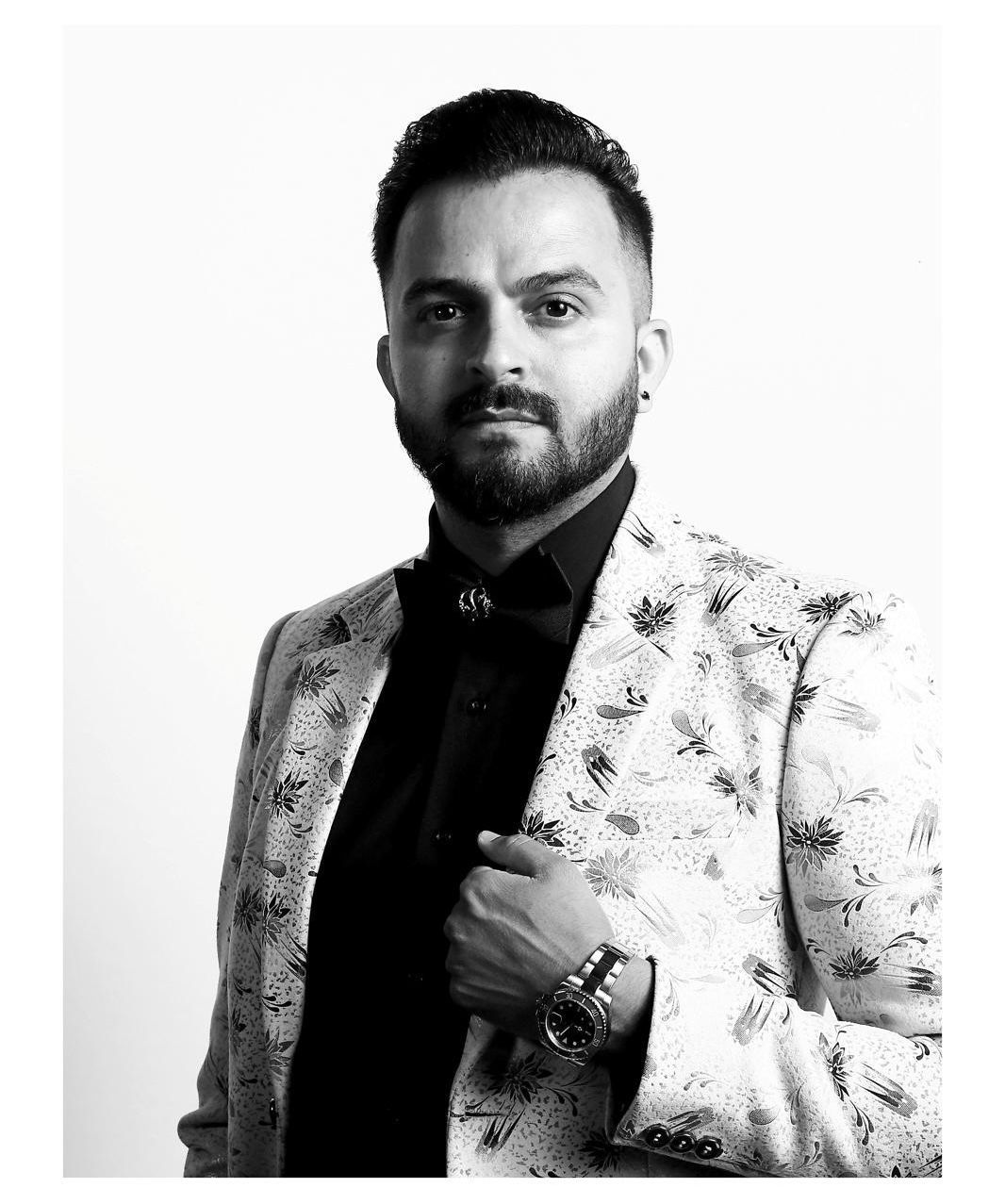 Man in patterned blazer, looking directly at the viewer. Black shirt, bow tie, watch. White background.