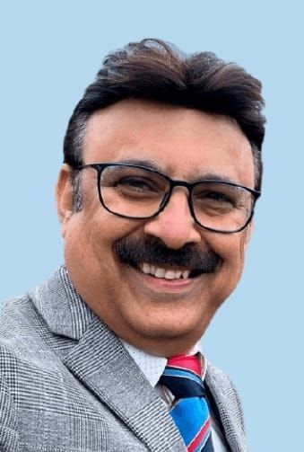 Smiling man with glasses, mustache, and tie, wearing a suit jacket against a blue backdrop.