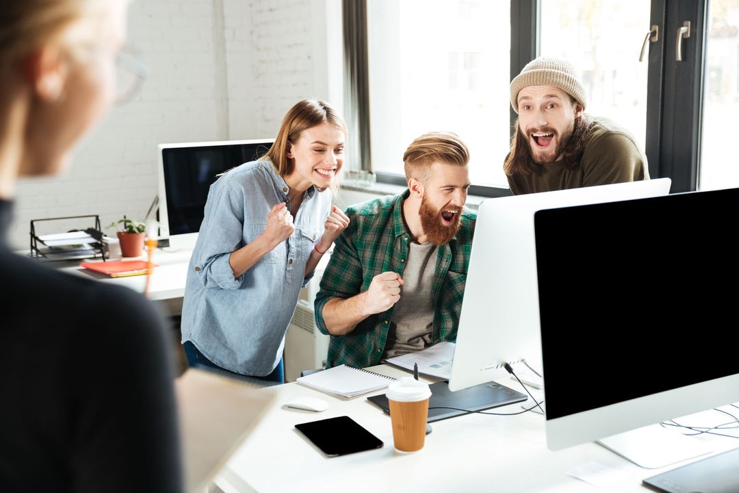 Excited office workers celebrating success in front of a computer; bright office setting.
