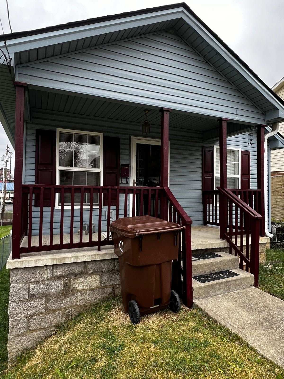 Small gray house with a front porch, brown railing, and a trash bin by the steps.