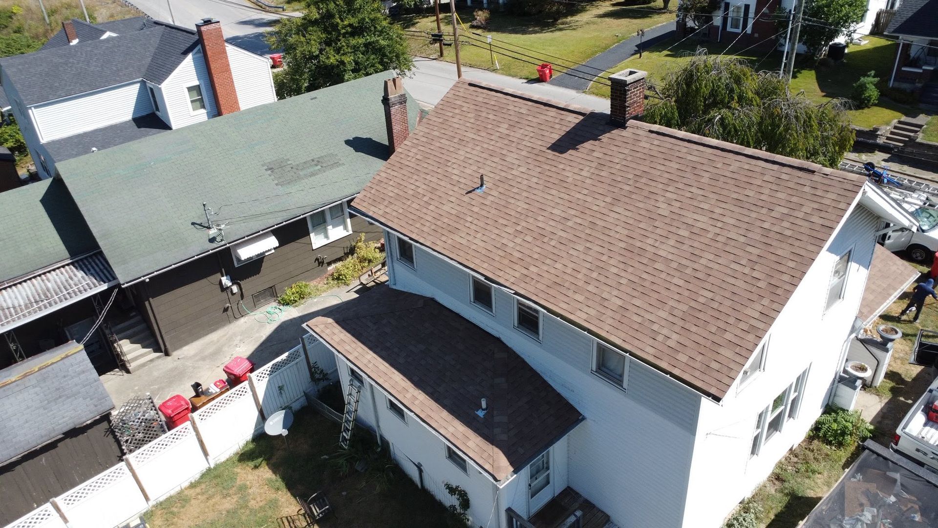 Aerial view of two houses with brown and green roofs, a white courtyard, and parked cars nearby.