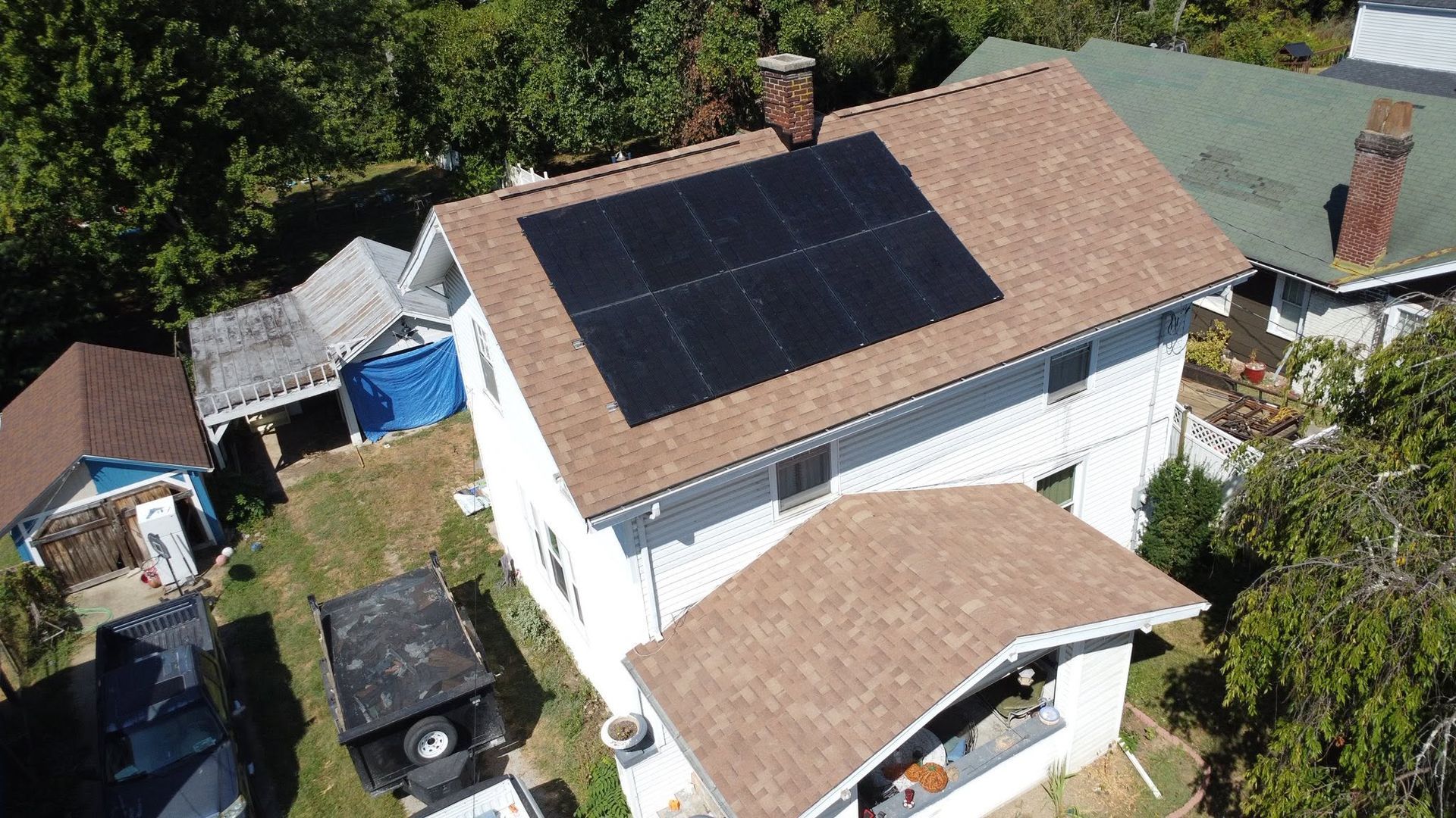 Aerial view of a white house with solar panels on the roof, surrounded by trees and neighboring homes.