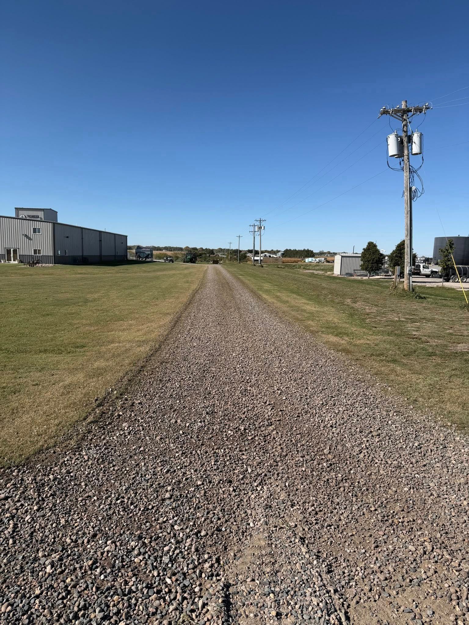 Gravel path between grassy fields, leading to buildings under a blue sky, power lines on the right.