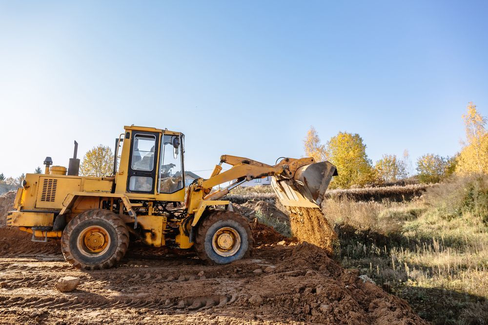 Yellow front-end loader dumping dirt onto a hillside under a blue sky.