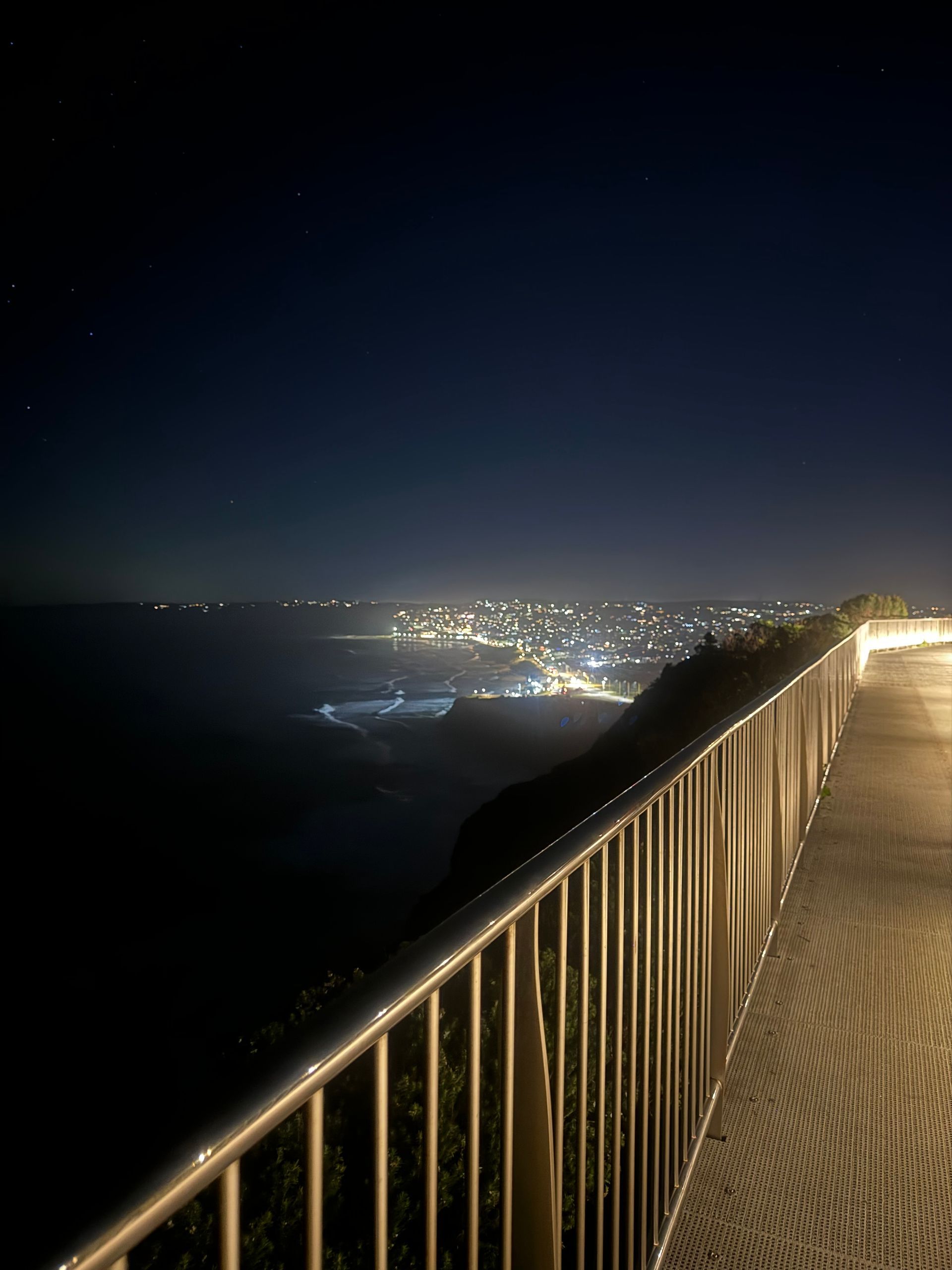 A lit walkway overlooks a dark coastline and a city glowing in the distance at night.