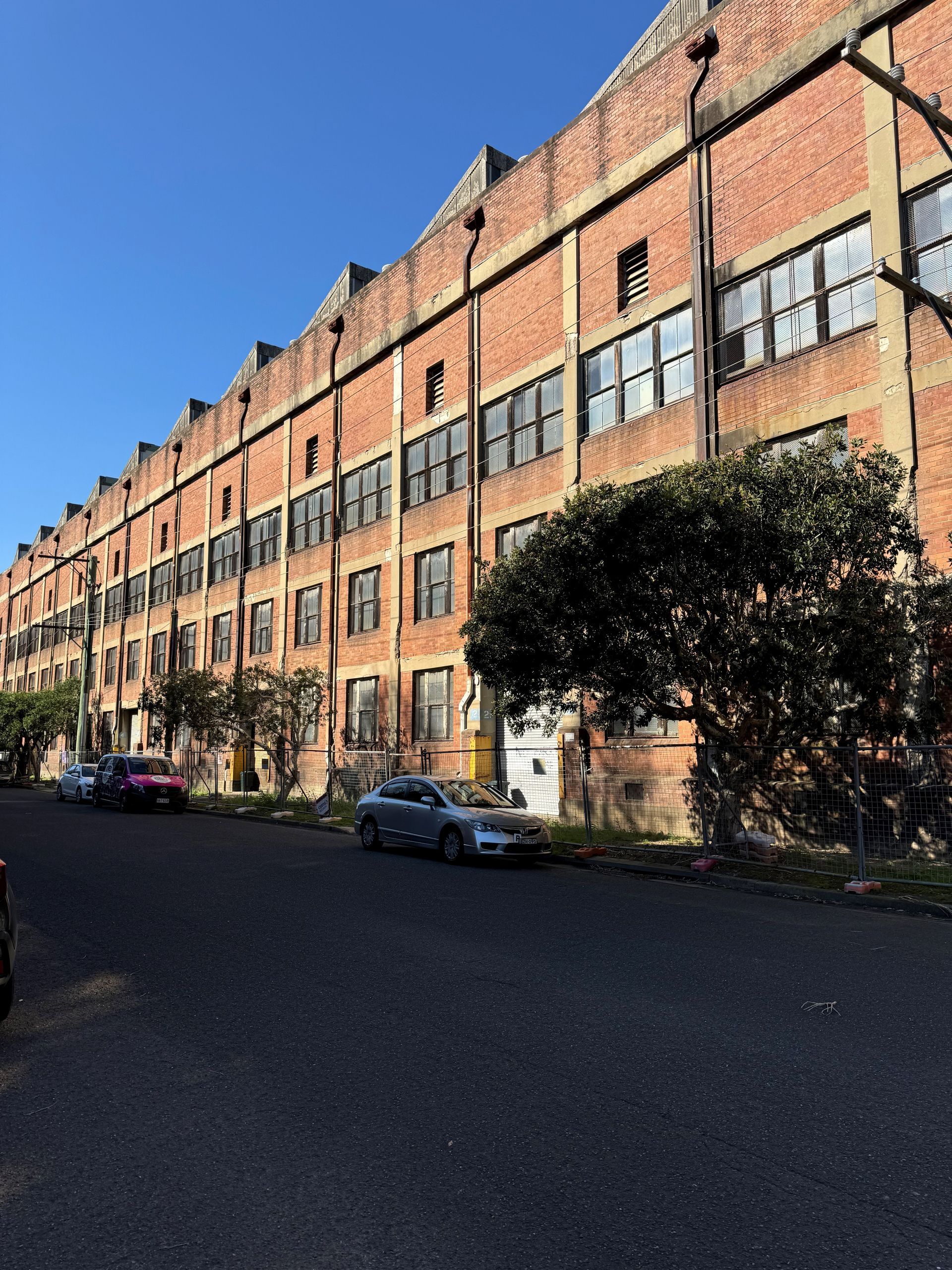 A long, red brick industrial building with many grid windows, parked cars, and trees on a sunny street.