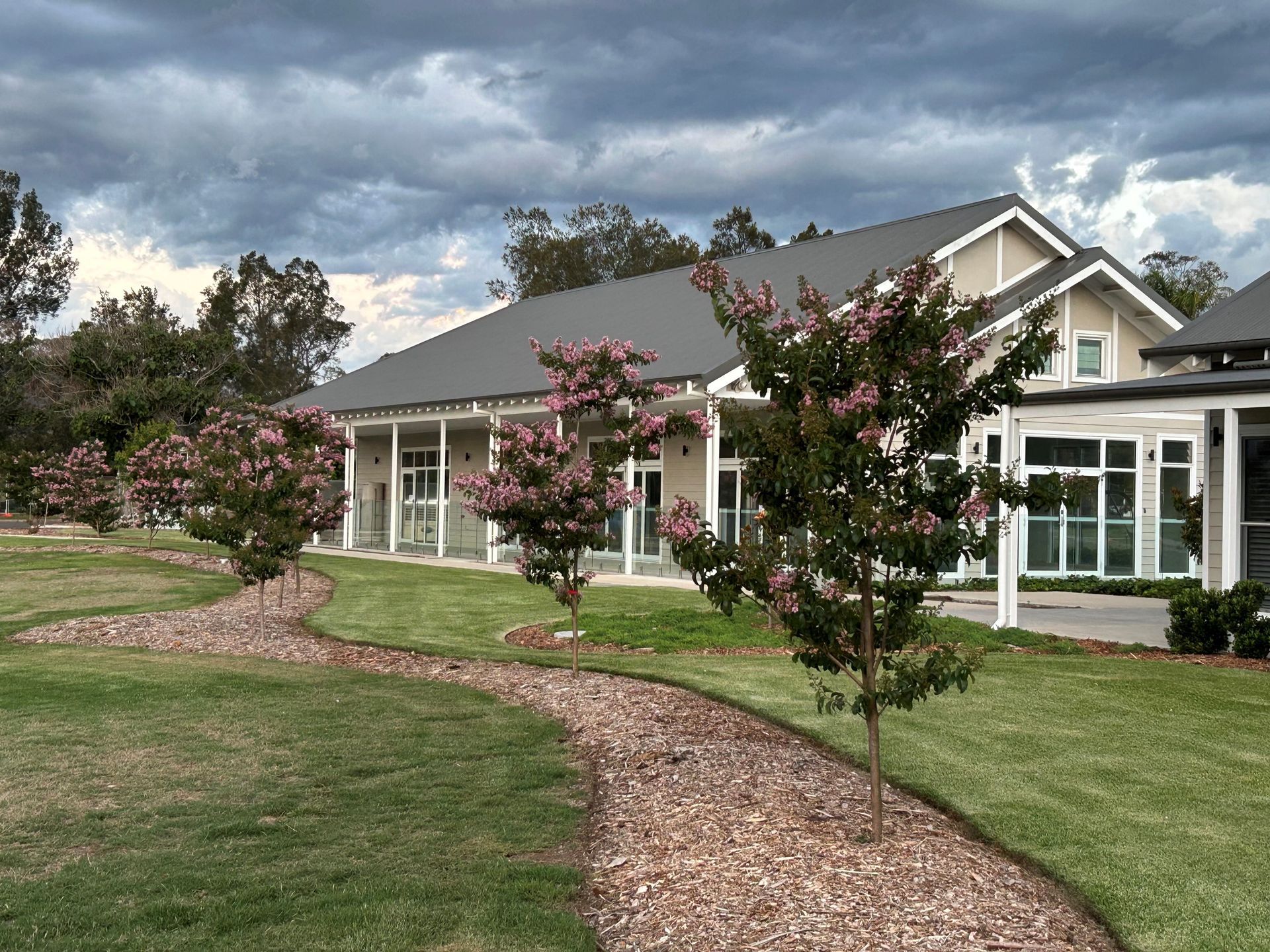 A beige building with a gray roof sits behind a curved gravel path lined with small flowering trees on a cloudy day.