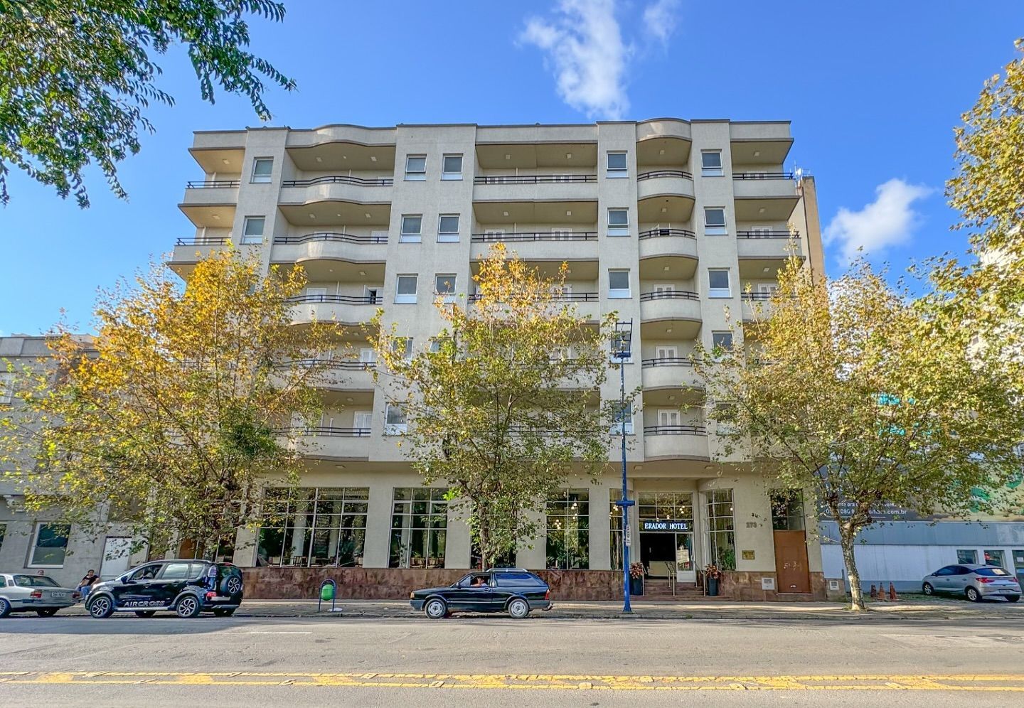Prédio de apartamentos com varandas, vitrines e carros estacionados em uma rua, sob um céu azul.imperador hotel