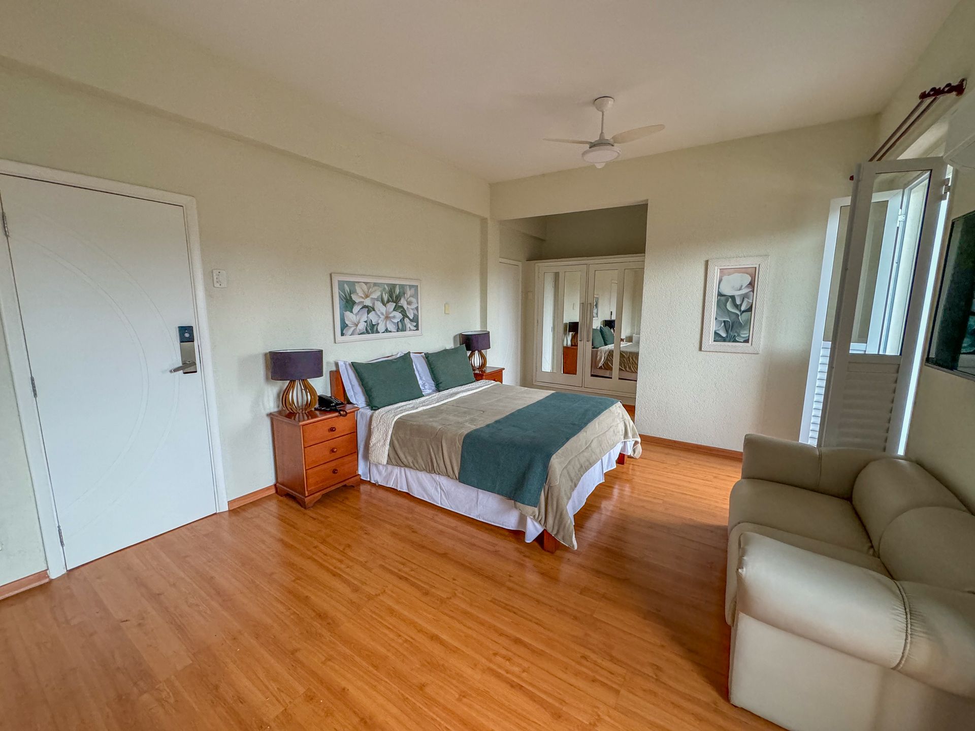 Quarto com piso de madeira, cama king size, sofá bege, porta e biombo decorativo.imperador hotel