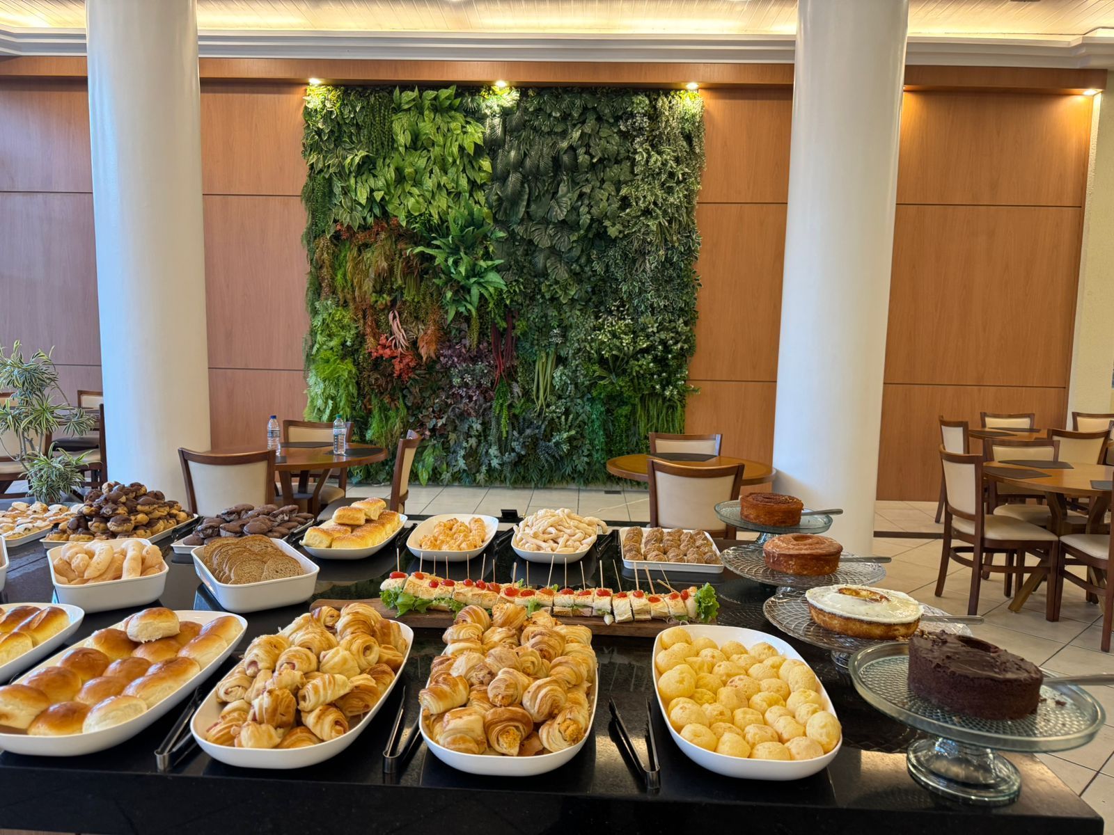 Mesa de bufê com diversos doces, bolos e pães. Parede verde ao fundo em uma sala iluminada.imperador hotel
