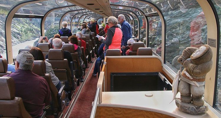 Interior of a glass-roofed train car with passengers; a figurine sits on a ledge.