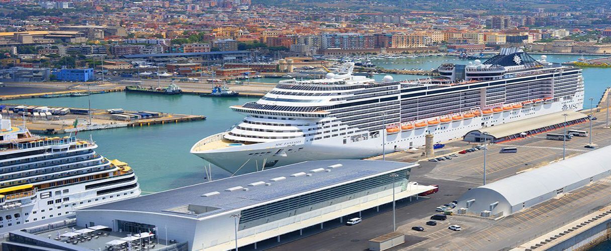 Large cruise ship docked in a harbor, with city buildings in the background. Sunny day.