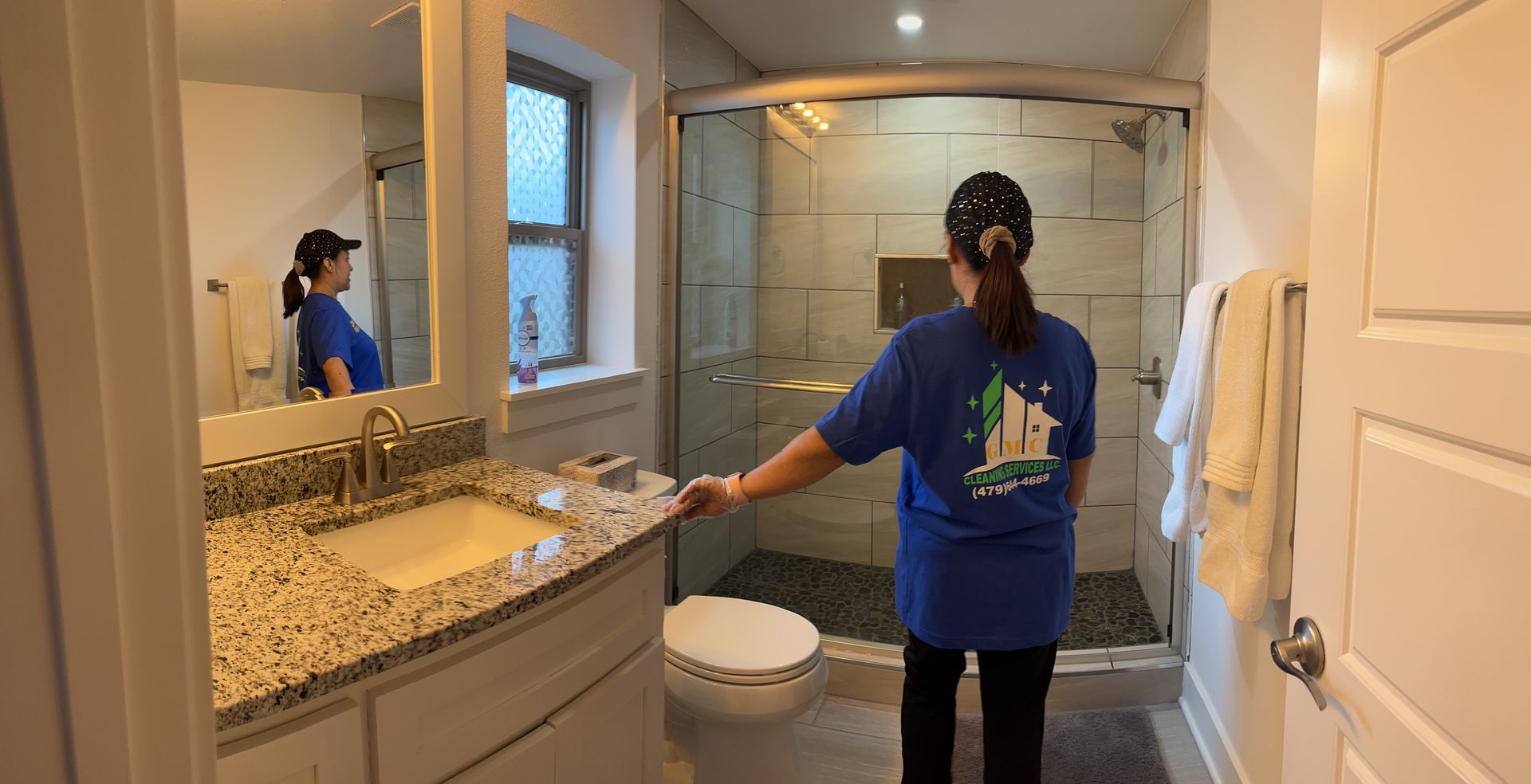 Woman in blue shirt stands in a bathroom with a glass shower and sink with a granite countertop.