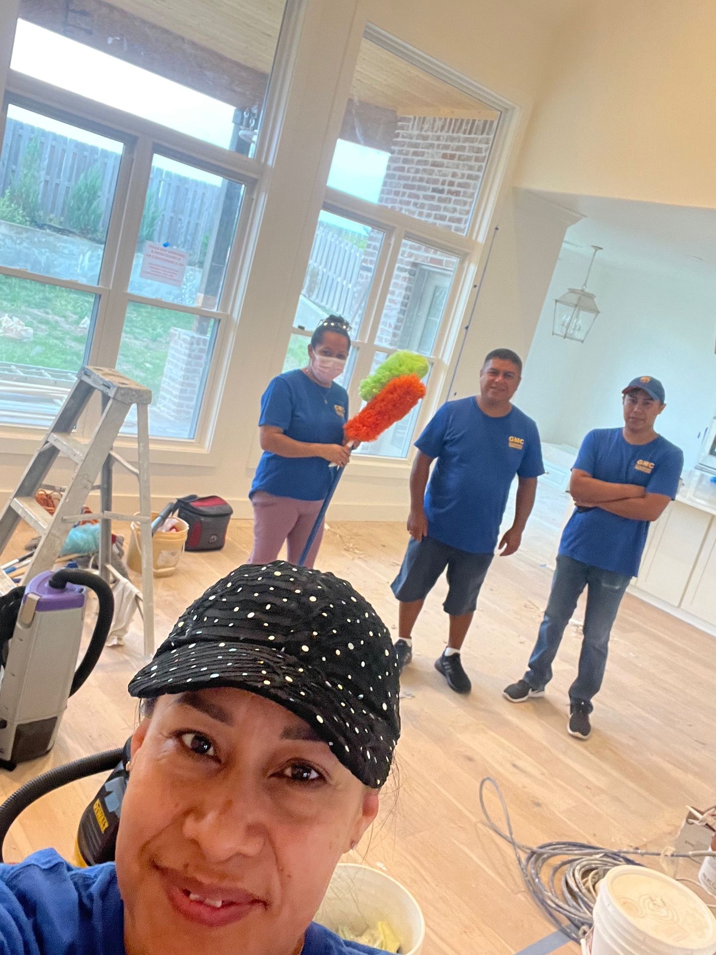 Four people cleaning a room with large windows and light wood floors. One person takes a selfie.