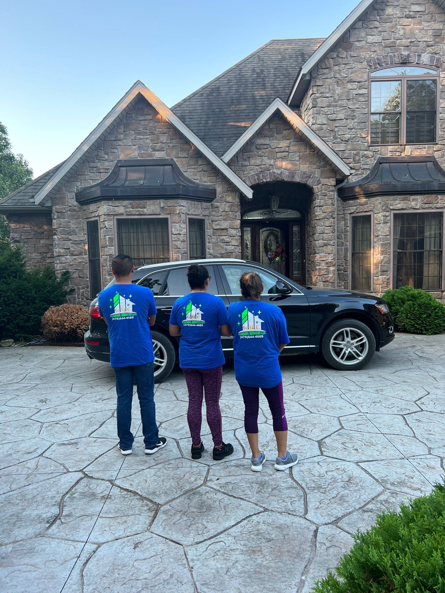 Three people in blue shirts stand in front of a large house and SUV, looking towards the entrance.