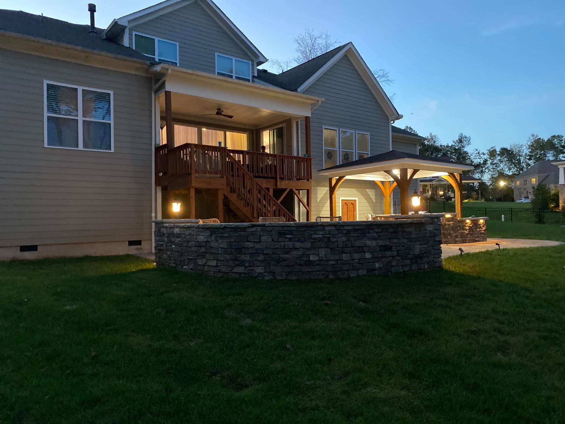 Backyard with house, deck, stone wall, and gazebo, illuminated at dusk.
