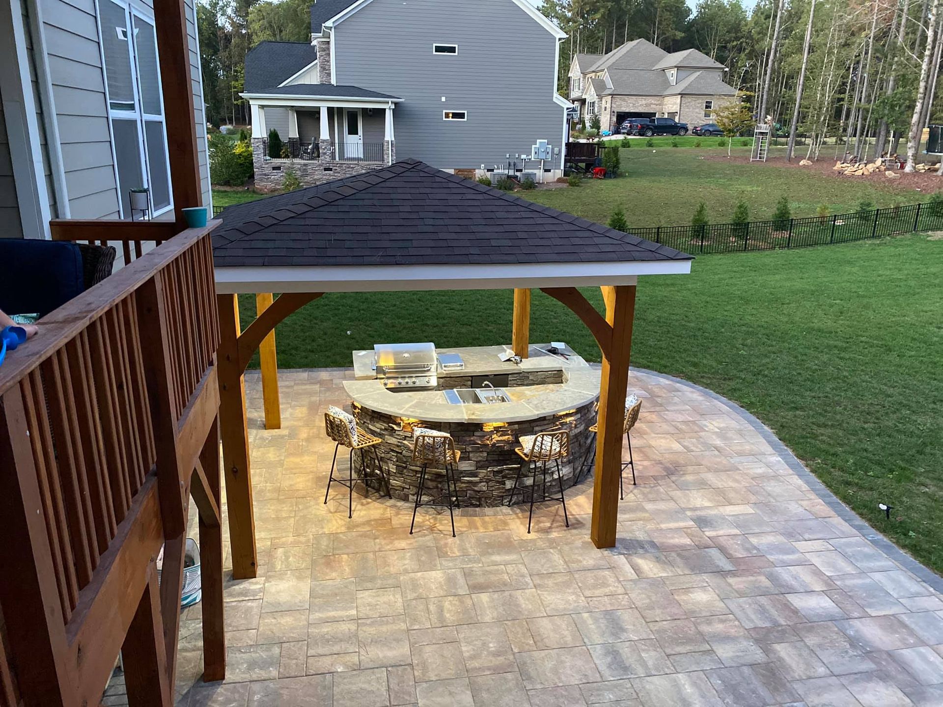 Outdoor patio with a gazebo over a stone fire pit, surrounded by chairs, and a grassy backyard.