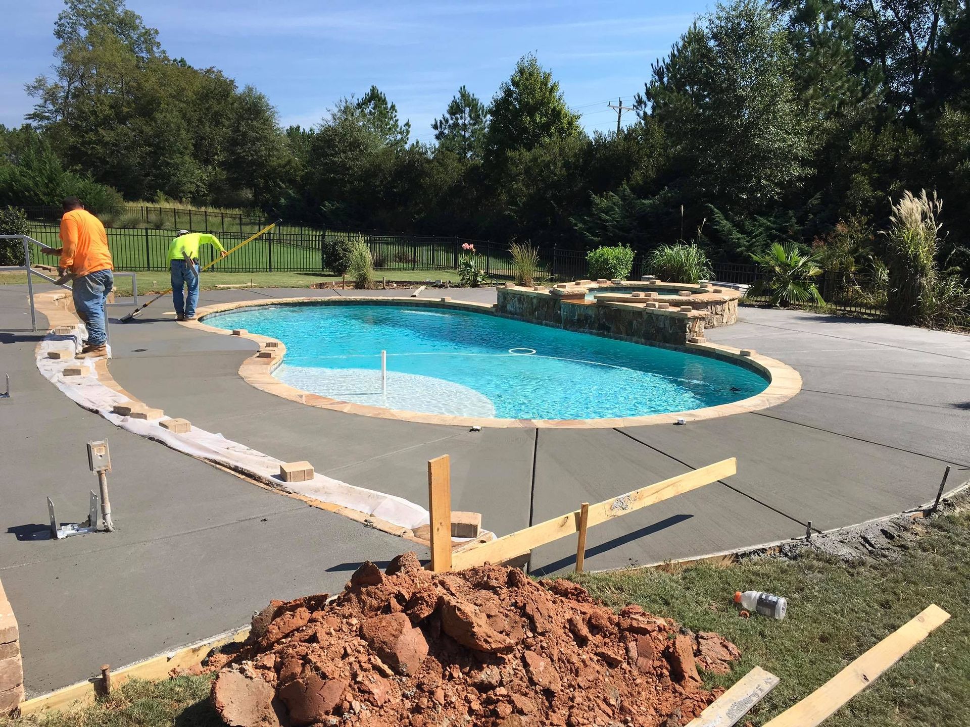 Workers pouring concrete around a completed pool. Green grass and trees in the background. Sunny day.