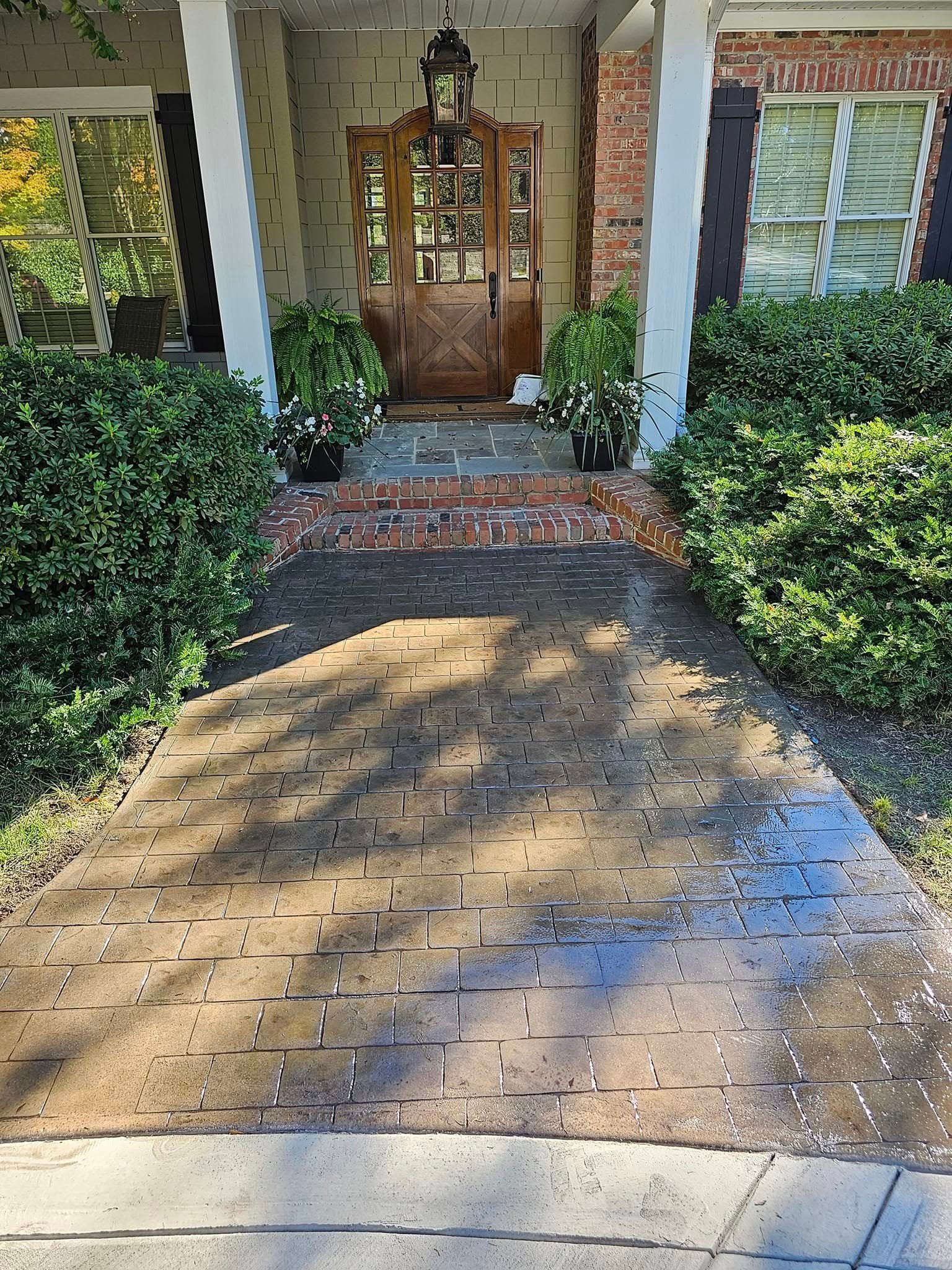 Brick pathway leads to a wooden door with brick steps; green bushes flank the path, a porch with white columns.
