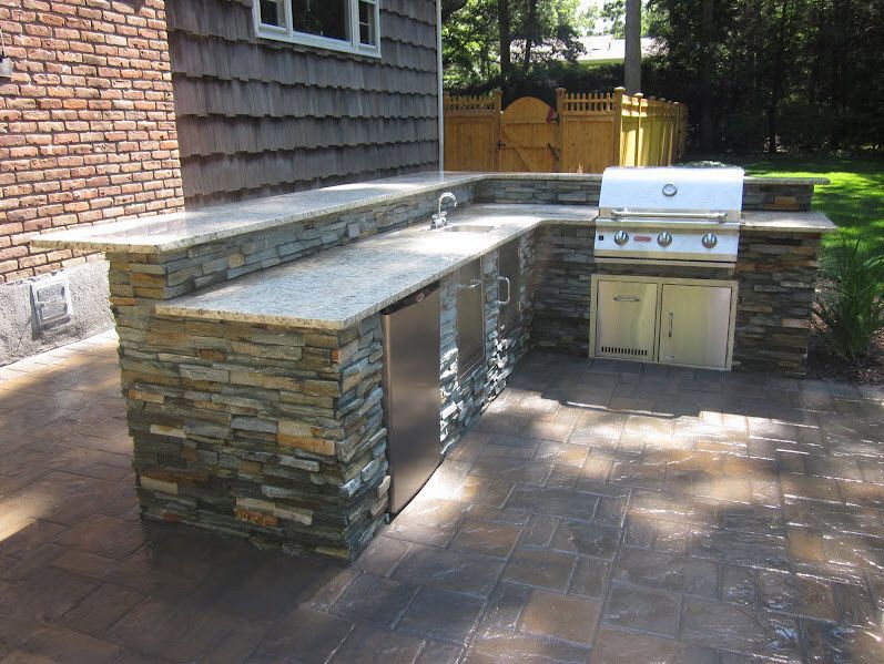 Outdoor kitchen with stone veneer and granite countertops. Features a grill, sink, and refrigerator.