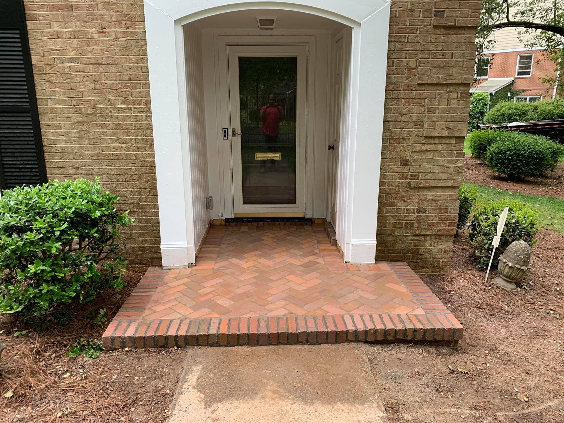 Brick entrance with glass door framed by white trim and flanked by bushes.