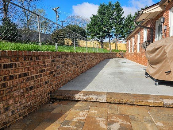 Brick retaining wall and patio next to a house with a concrete driveway and chain link fence.