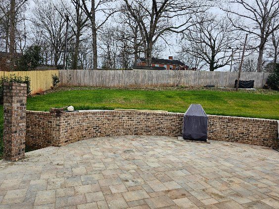 A stone-faced retaining wall with a covered grill on a brick patio, overlooking a grassy hill and trees.