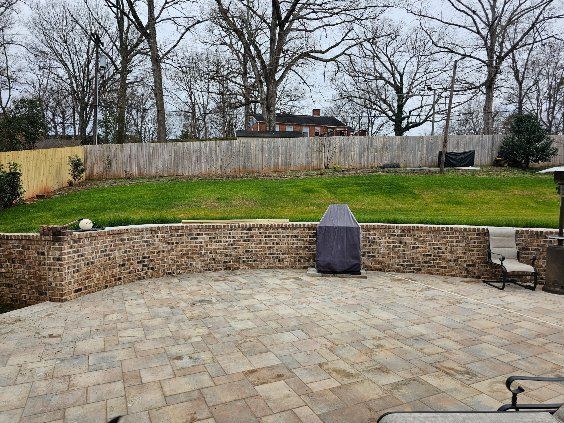 A paved patio with a brick retaining wall and a grill, green grass, and a wooden fence. Overcast day.