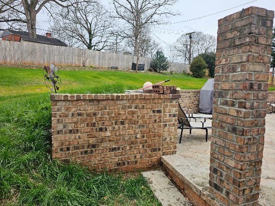 Brick retaining wall in a backyard with patio and wooden fence. Overcast sky and green grass.
