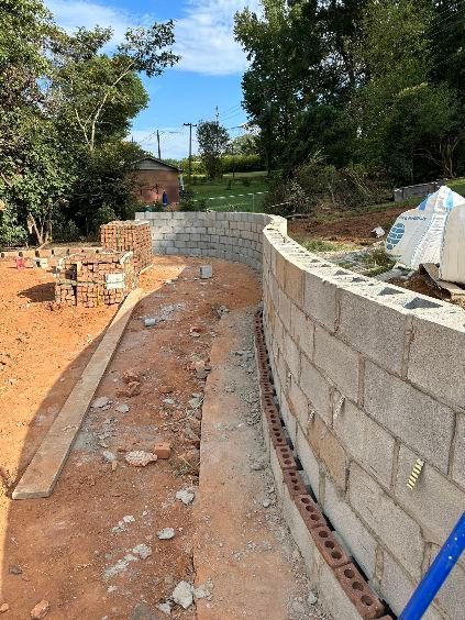 Construction of a curved retaining wall. Blocks are grey, ground is red clay, blue sky with trees in the background.