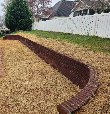 A low, curved, brick retaining wall on a grass and mulch-covered slope, with a white picket fence in the background.
