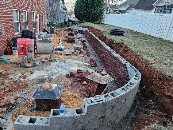 Brick retaining wall under construction in a yard. Cement blocks form the base, with a brick facade rising above.
