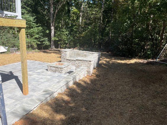 Stone patio with fire pit and built-in stone seating area, surrounded by brown grass and trees.