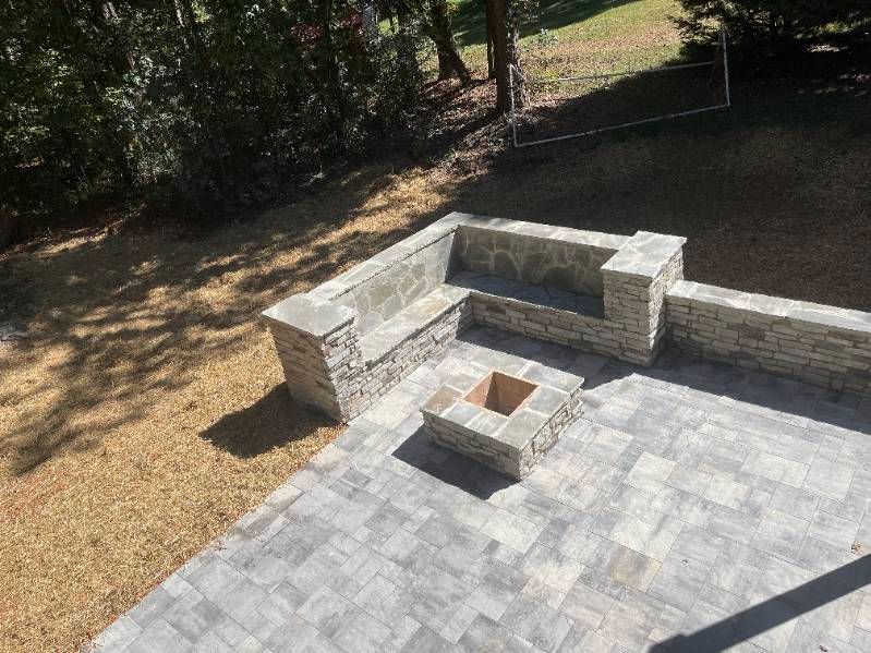 Stone patio with built-in seating, a fire pit, and surrounding yard under sunlight.