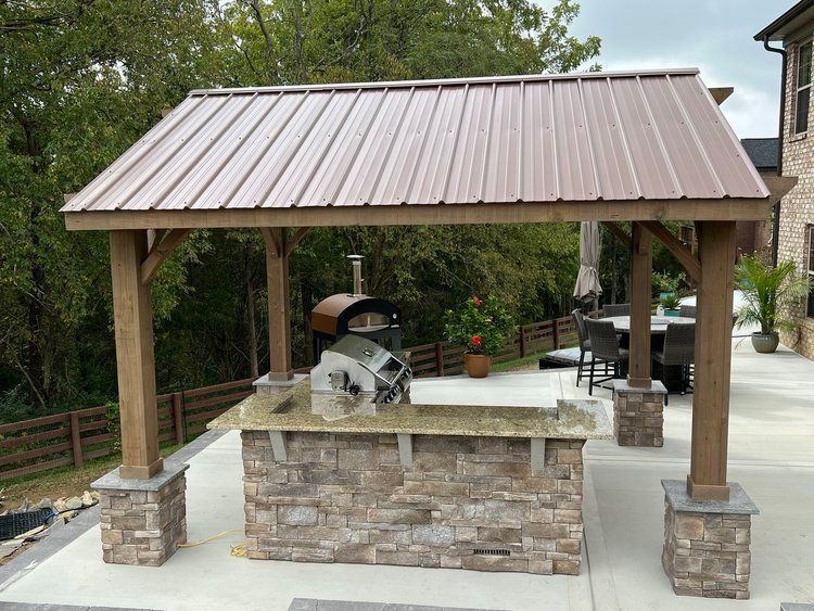 Outdoor kitchen with brown metal roof, stone facade, and pizza oven.