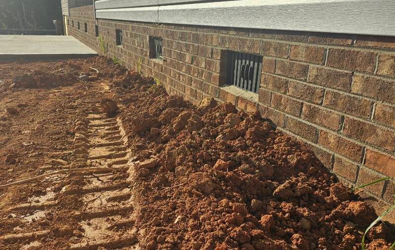 Trench of red clay next to a brick building with small window wells and a concrete sidewalk.