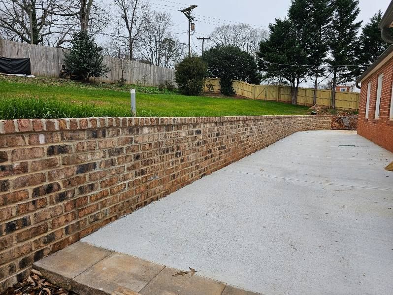 Brick retaining wall bordering a concrete patio, with a grassy hill and trees in the background.
