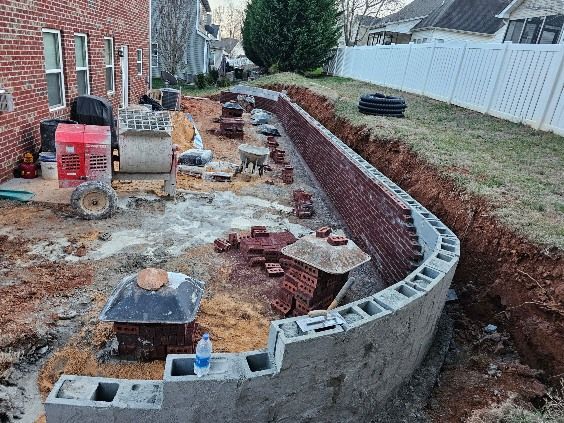 Construction site: brick retaining wall under construction next to a house and fence.