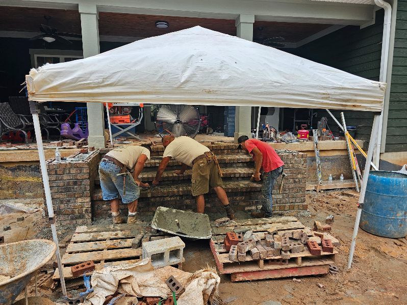 Three workers building stone steps under a white canopy on a porch; construction materials are visible.