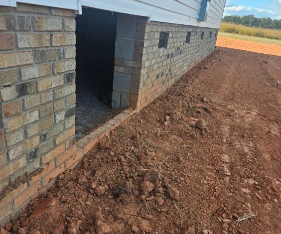 Brick foundation of a building with a crawl space opening, and dirt ground.