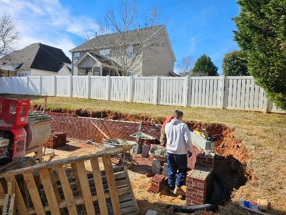 Construction site with a person carrying bricks. A brick retaining wall is being built in a backyard with a white fence.