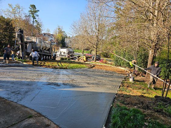 Workers pouring and smoothing fresh concrete driveway with trucks and equipment in wooded setting.
