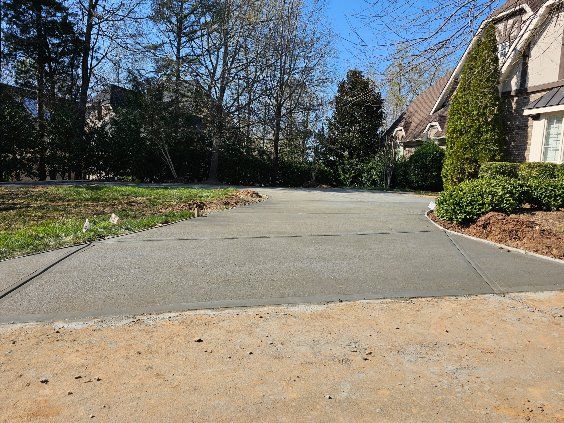 Concrete driveway leading to a house, surrounded by grass, trees, and landscaping.