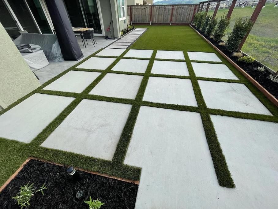 Patio with square concrete pavers and artificial grass. Outdoor setting with a fence and landscaping.