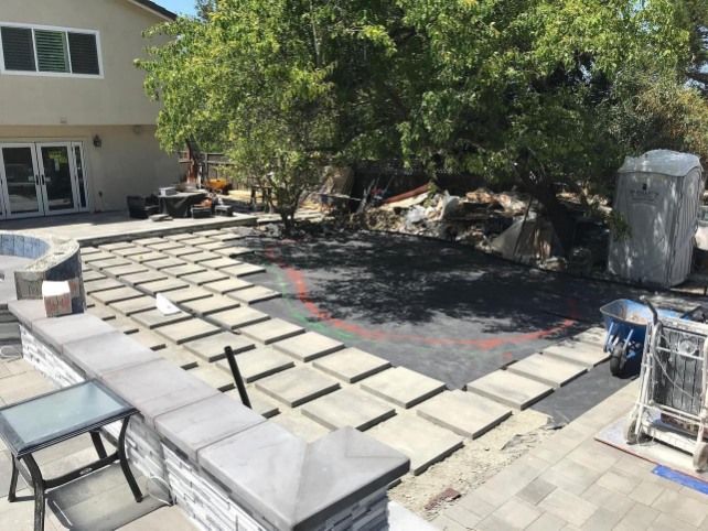 Patio construction: paving stones laid around a partially-excavated area with a tree and a retaining wall.
