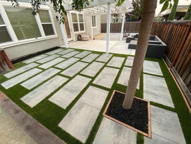 Backyard patio with gray rectangular pavers set in green turf around a tree and a seating area.